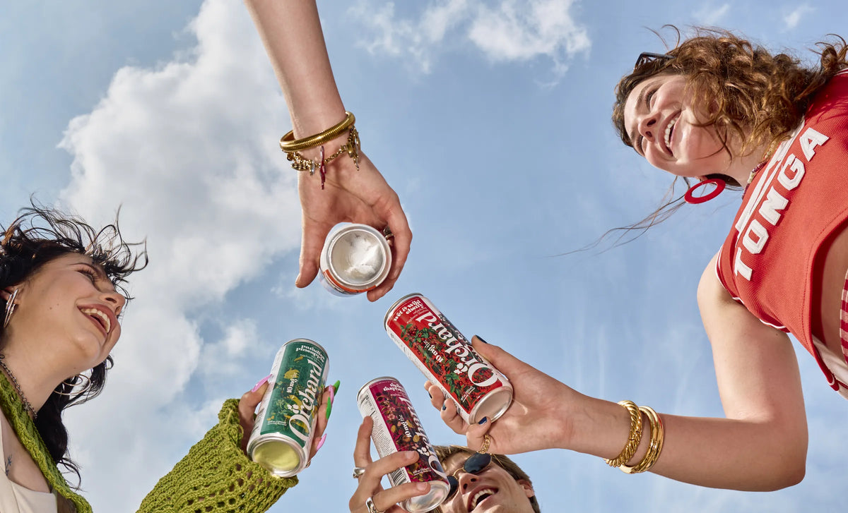 four friends clinking Orchard cans against a blue sky